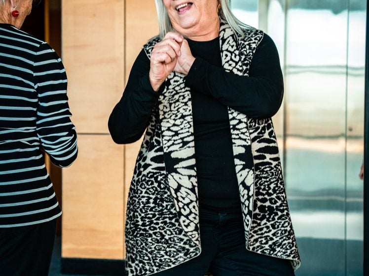 Image of An older woman with long grey hair, glasses, and a patterned black-and-white waistcoat smiles and gestures while talking to another person in a striped shirt, standing indoors near a lift.