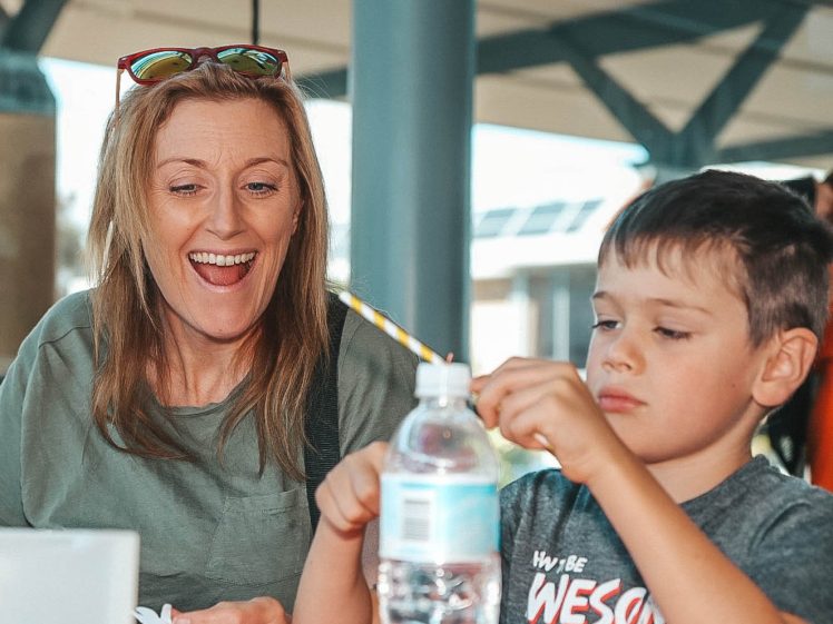 Image of A smiling woman watches as a young boy concentrates on inserting a striped straw into a plastic water bottle at a table indoors.