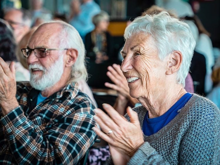 Image of Two older adults with grey hair, wearing glasses and casual jumpers, sit side by side, smiling and clapping in a lively, crowded indoor setting. The focus is on their joyful expressions.
