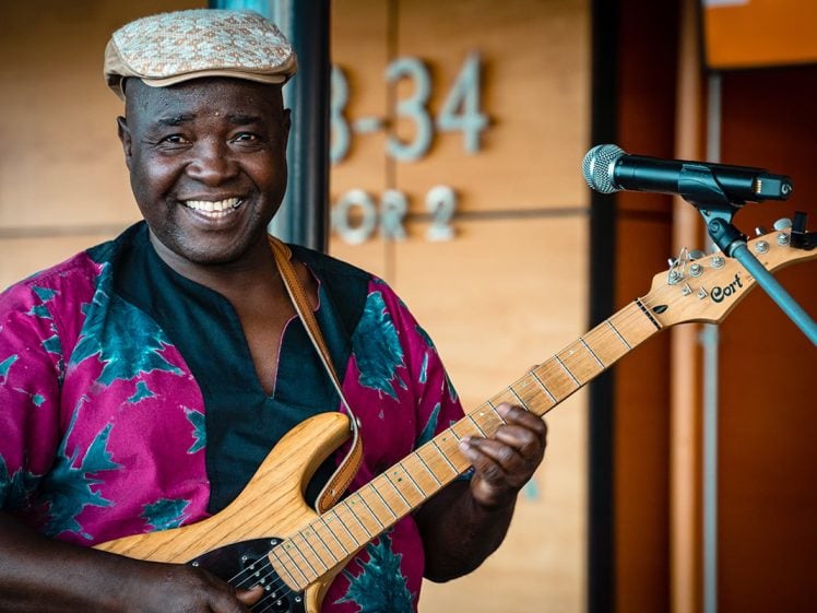 Image of A smiling man wearing a patterned shirt and cap plays an electric guitar next to a microphone stand, standing in front of a wooden wall with numbers in the background.