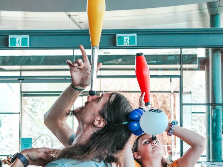 Image of Two people balance juggling clubs on their faces indoors. The person in front balances a yellow club on their chin and has long hair, whilst the person behind, wearing a balloon hat, balances a red club on their forehead.