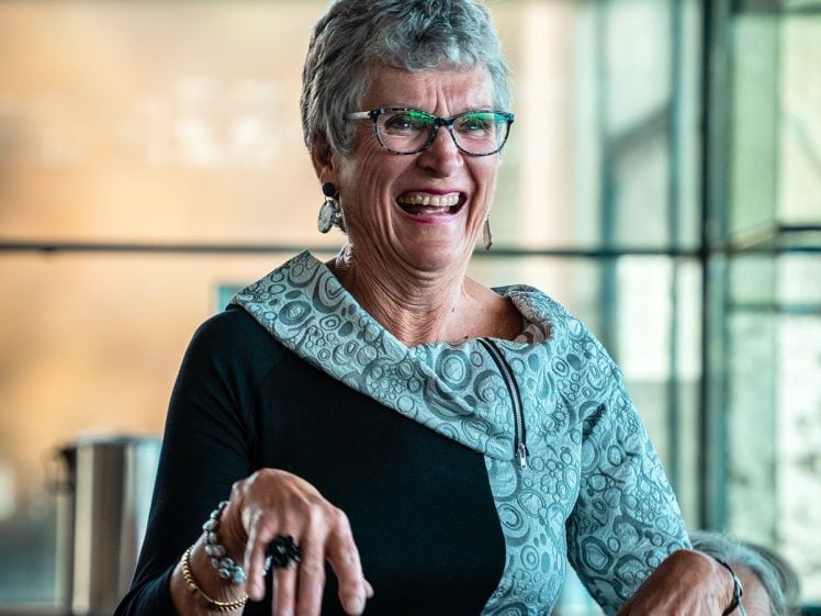 Image of An older woman with short grey hair and glasses smiles broadly while standing indoors. She is wearing a black and grey patterned dress and jewellery, and her hands are playfully raised in front of her.