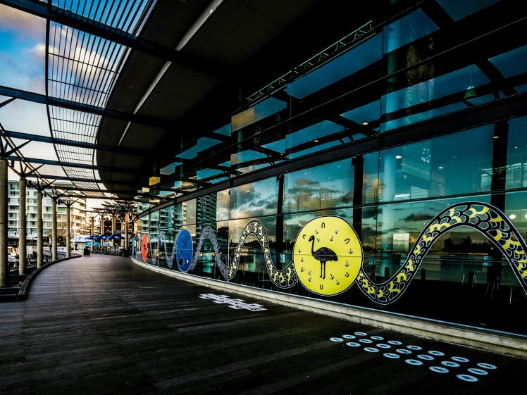 Image of A modern waterfront walkway with a large glass building featuring colourful circular art and a yellow snake-like mural. Reflections of buildings and the sky are visible in the glass panels.