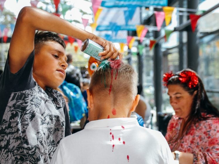 Image of A boy sprays colourful hair paint on another boy’s head at a festive indoor event with bunting flags. A woman in red headband ears sits nearby, focused on an activity.
