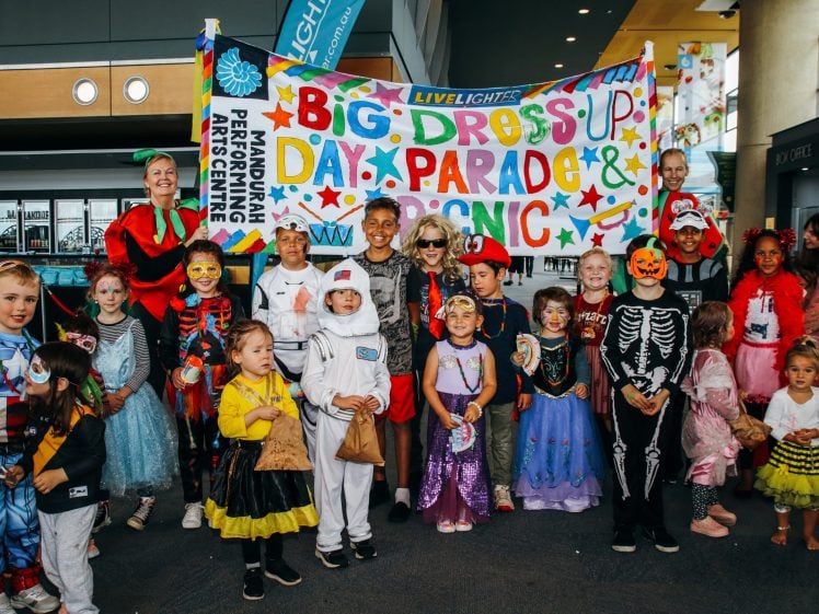 Image of A group of children and adults in colourful costumes stand in front of a Big Dress Up Day Parade & Picnic banner indoors, smiling and posing for the camera.