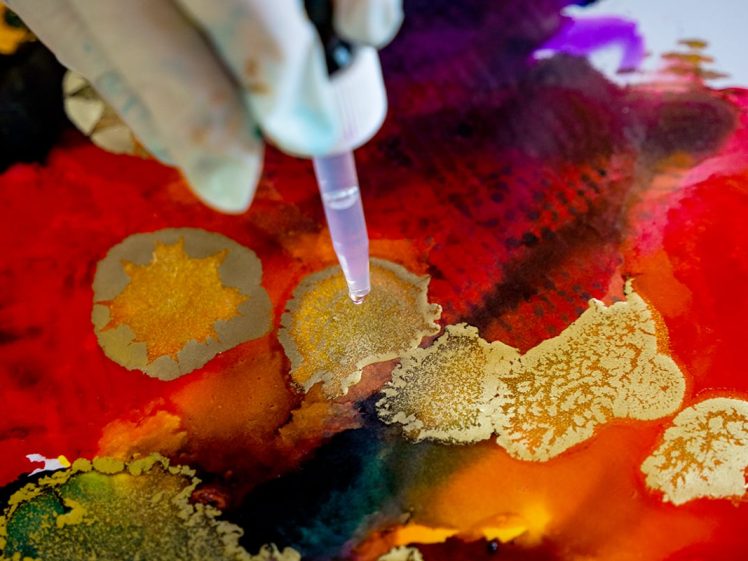 Image of A gloved hand uses a dropper to add liquid to vibrant watercolour and gold ink patterns on paper, blending red, orange, and purple hues with metallic accents.