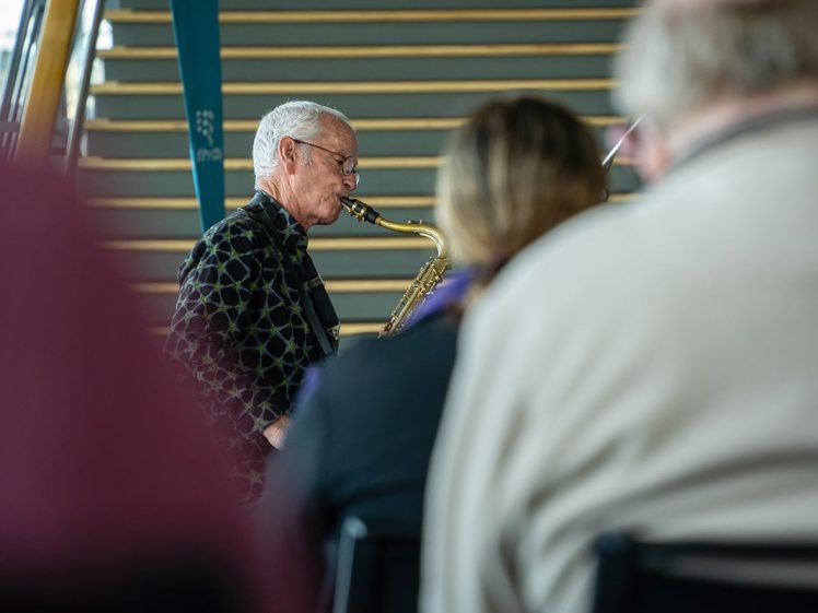 Image of An older man wearing glasses and a patterned jacket plays a saxophone indoors, with blurred people in the foreground and a staircase in the background.