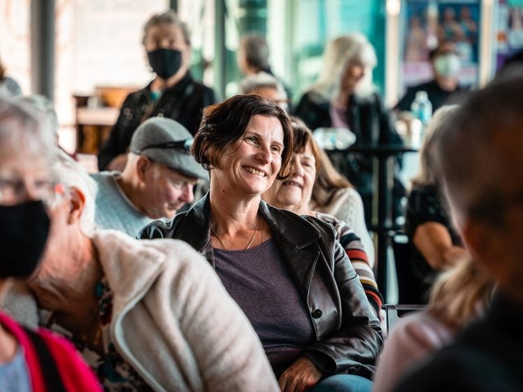 Image of A woman in a leather jacket smiles whilst sitting among a group of people, some wearing face masks, at an indoor event. The atmosphere appears friendly and engaged.