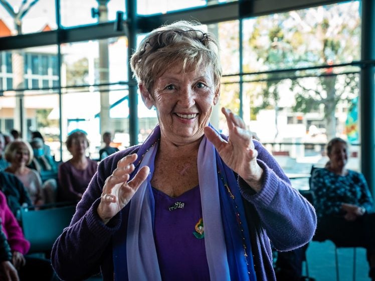 Image of An older woman in a purple outfit smiles and gestures with her hands whilst standing indoors, with a seated audience and large windows behind her. The atmosphere appears bright and cheerful.