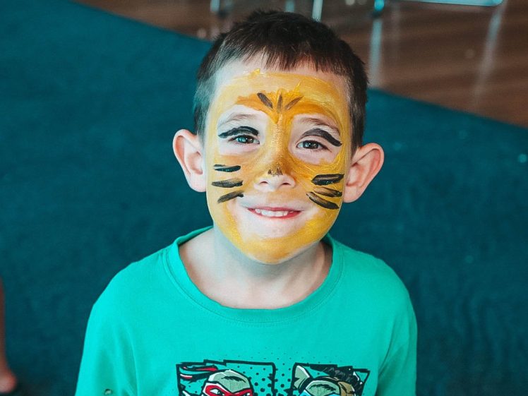 Image of A young boy with short brown hair smiles, wearing a green Teenage Mutant Ninja Turtles shirt and face paint resembling a tiger, with yellow colouring and black stripes. He stands indoors on a teal carpet.