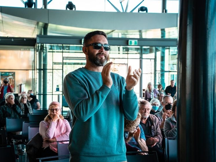 Image of A man in sunglasses and a blue jumper stands clapping in a modern, glass-walled room filled with seated people, some of whom are also clapping.