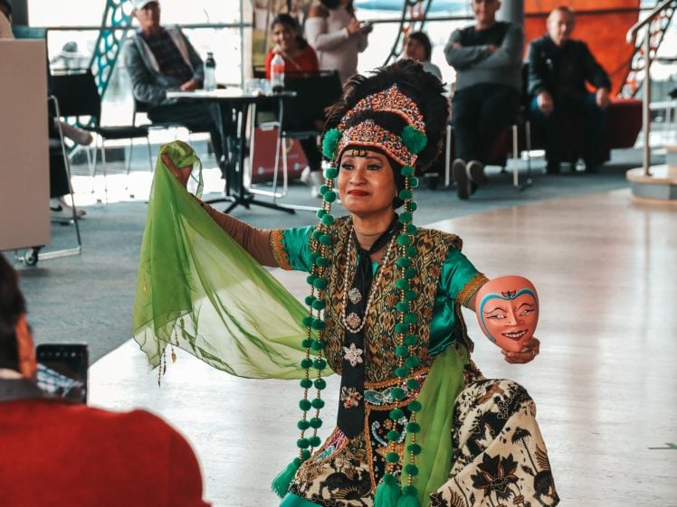 Image of A performer in ornate traditional attire and headdress holds a green scarf and a painted mask, kneeling on the floor during a cultural performance, with an audience seated in the background.