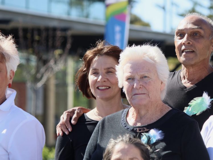 Image of A group of older adults stands outdoors, smiling and posing for a photo. They are wearing black or white clothing and have feather pins on their outfits. A blurred building and banner are visible in the background.