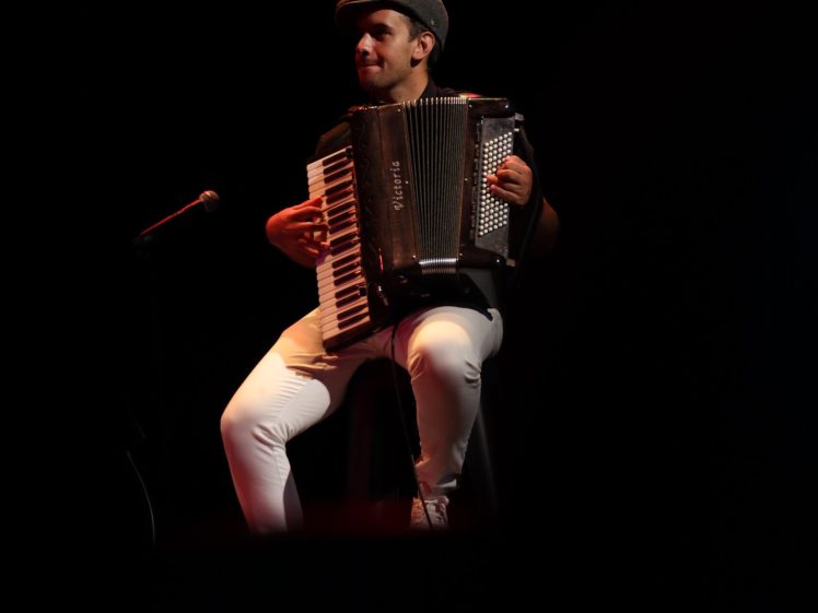 Image of A person wearing a cap and white trousers plays an accordion while seated on a stool under a spotlight, with a microphone stand nearby, against a dark background.