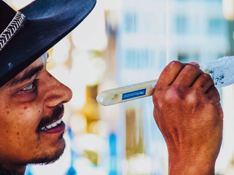 Image of A person wearing a black hat and smiling paints a surface with a white paintbrush, focusing intently on their work. The background is blurred, highlighting the painter and their hand.