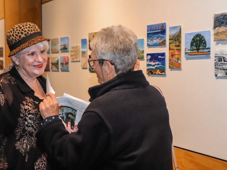 Image of Two women converse in front of a gallery wall displaying small, colourful landscape paintings. One woman wears a leopard-print hat and patterned black top, while the other holds a book and wears glasses and a dark jacket.