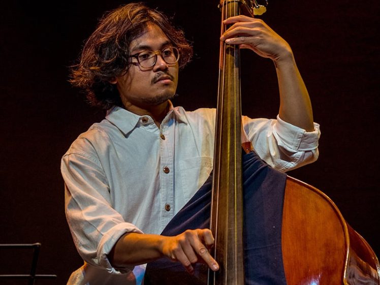 Image of A musician with medium-length dark hair and glasses plays a double bass on stage, wearing a white button-up shirt, with a dark background highlighting the performance.
