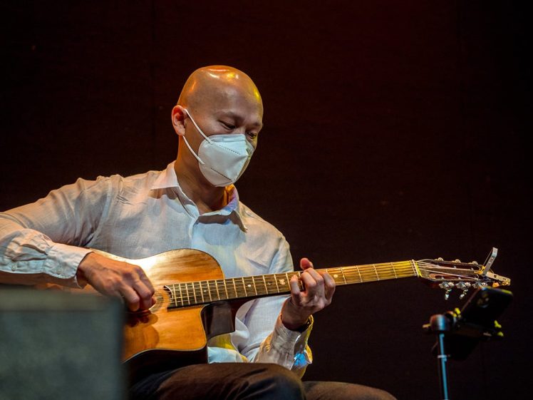 Image of A person wearing a white shirt and a face mask plays an acoustic guitar on stage against a dark background.