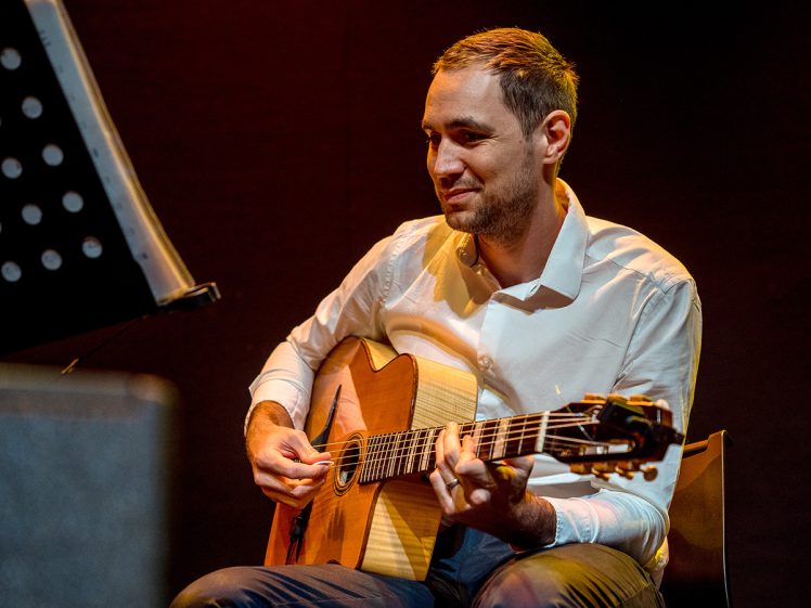 Image of A man in a white shirt sits and plays an acoustic guitar, looking at sheet music on a stand. The background is dark and he appears focused and contented.