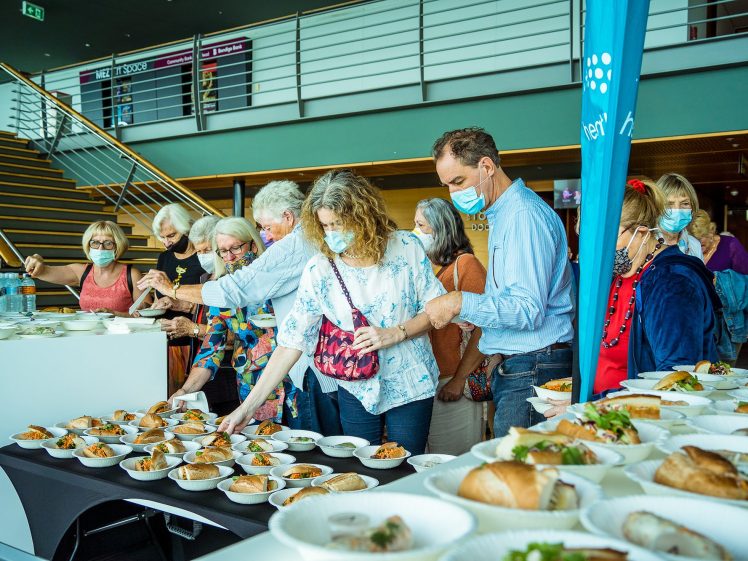 Image of A group of people wearing face masks stands in queue at a buffet table with assorted food dishes, including sandwiches and salads, in a modern indoor setting.