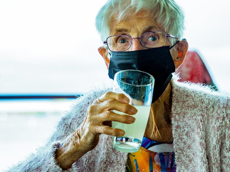 Image of An elderly person with white hair and glasses wears a black face mask and holds a glass of light yellow drink. They are seated indoors, wearing a colourful shirt and a light pink cardigan.