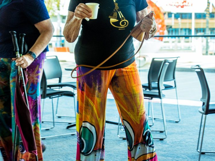 Image of An older woman wearing a black mask, black shirt, and colourful, wide-legged trousers holds a coffee cup and a hula hoop indoors. Another woman with walking sticks stands nearby. Rows of empty chairs are in the background.