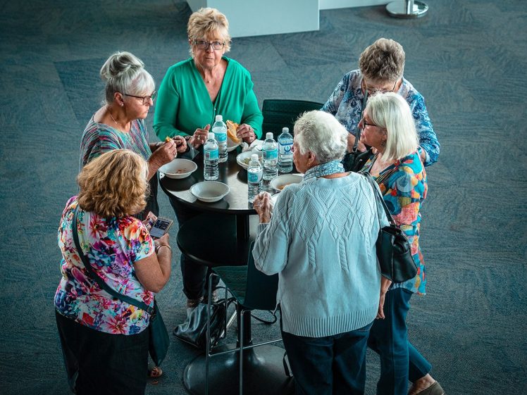 Image of Six older women stand around a small round table with drinks and snacks, chatting and smiling in a bright indoor setting. Some are wearing glasses and colourful clothing.