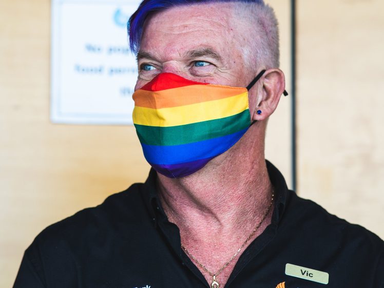 Image of A person with short blue hair wears a rainbow pride mask and a black shirt with volunteer badges. The background is blurred and light-coloured.