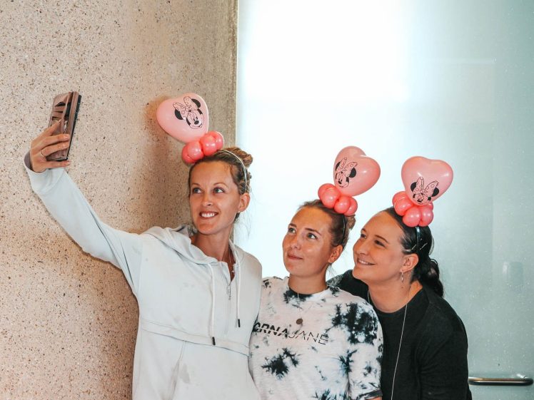 Image of Three women wearing heart-shaped Minnie Mouse balloon headbands smile and pose for a selfie together against a light-coloured wall.