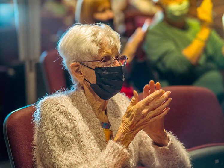 Image of An elderly woman with white hair, glasses, and a black face mask sits in an auditorium chair, clapping her hands. She wears a fluffy light pink jumper. Other masked people are seated in the background.