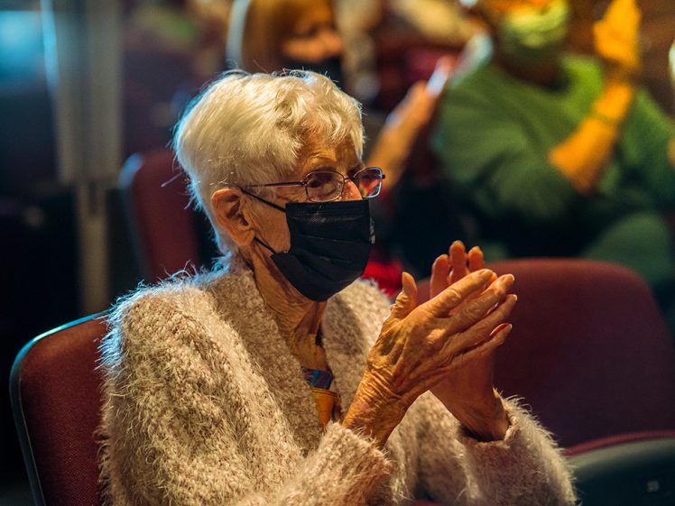 Image of An elderly woman with white hair wearing glasses and a black face mask is seated and applauding in an auditorium. Other masked people are visible in the blurred background.