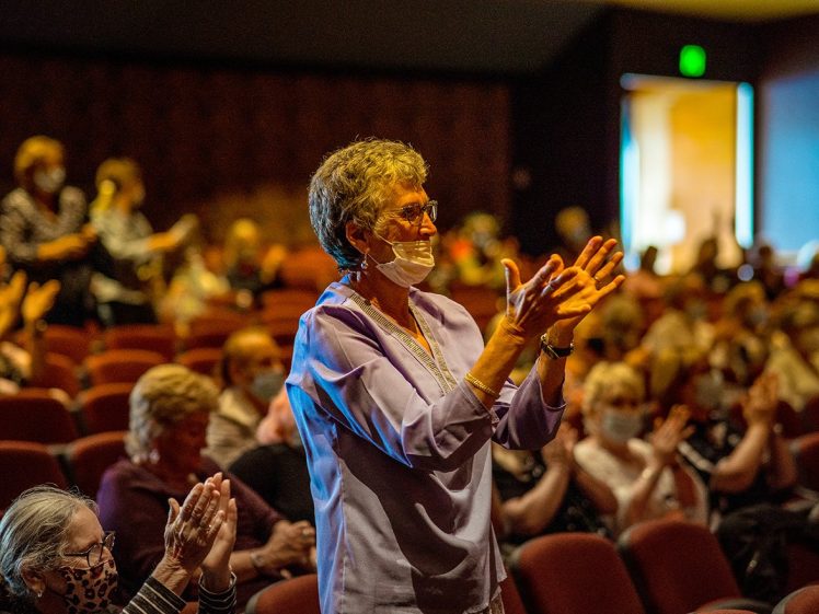 Image of An older woman wearing glasses and a mask stands and applauds in a theatre filled with seated people, some also clapping. Warm lighting highlights the scene.