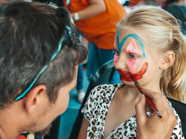 Image of A man paints a colourful butterfly and red smile on a young girl’s face at a face-painting event. The girl sits still in a chair, wearing a leopard print top, with people visible in the blurred background.