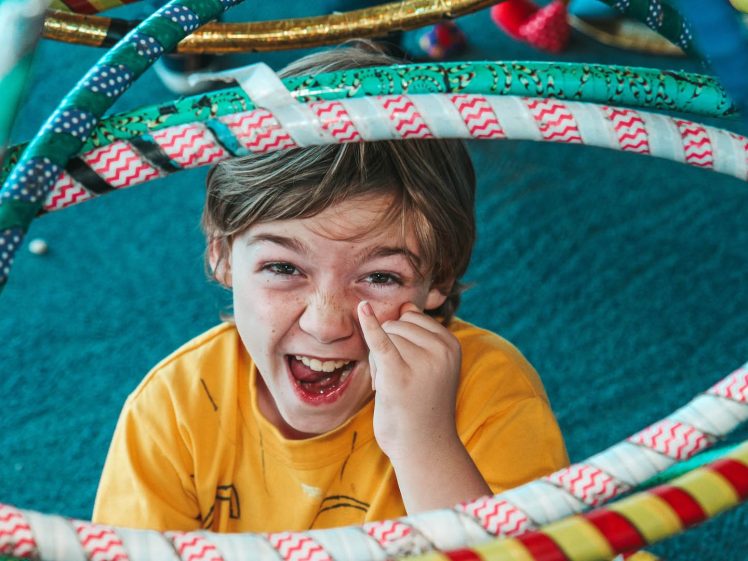 Image of A young boy in a yellow shirt smiles and laughs while looking up through colourful hula hoops, with one hand playfully touching his face. The background is blue-green carpeting.