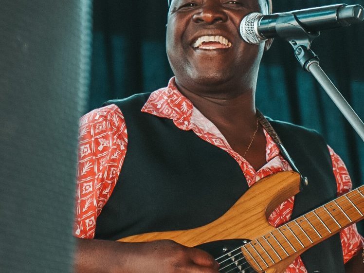 Image of A smiling man wearing a red patterned shirt, black waistcoat, and cap plays an electric guitar and sings into a microphone on stage.