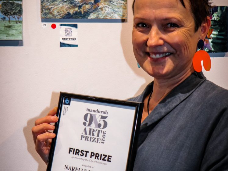 Image of A person smiling and holding a framed certificate for First Prize at the Mandurah 95 Art Prize. Artwork is displayed on the wall behind them.