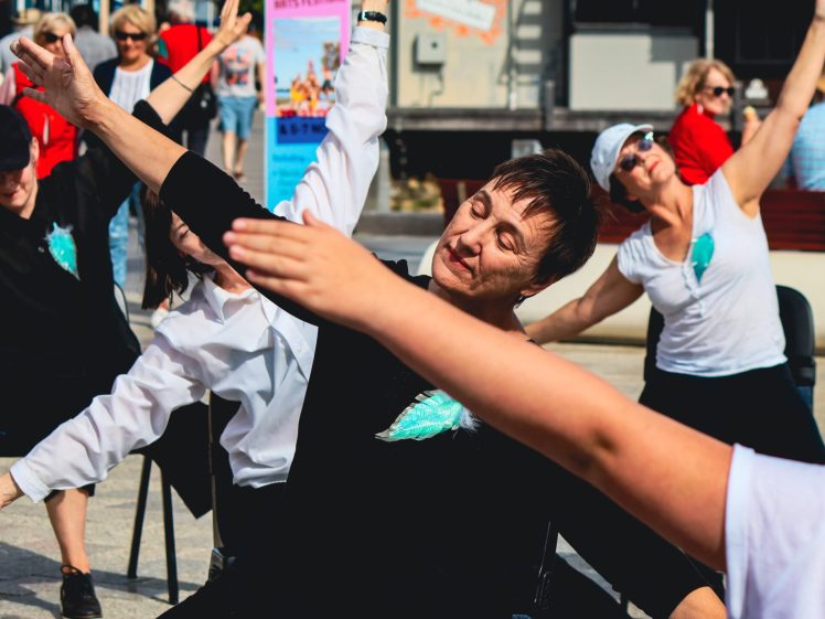 Image of A group of adults, mostly women, sit outdoors on chairs performing synchronised stretching exercises, extending their arms and leaning to the side. They appear to be enjoying the physical activity on a sunny day.
