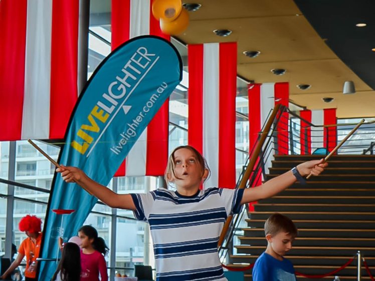 Image of A child in a striped shirt skilfully spins a diabolo with two sticks indoors. Red and white banners hang from the ceiling, and other children and adults are in the background near stairs.