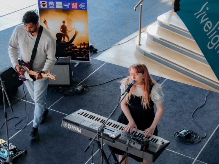 Image of A woman sings and plays a Yamaha keyboard while a man plays electric guitar on stage. There are microphones, amplifiers, and a music poster in the background. The scene is indoors, with blue carpeting and steps.