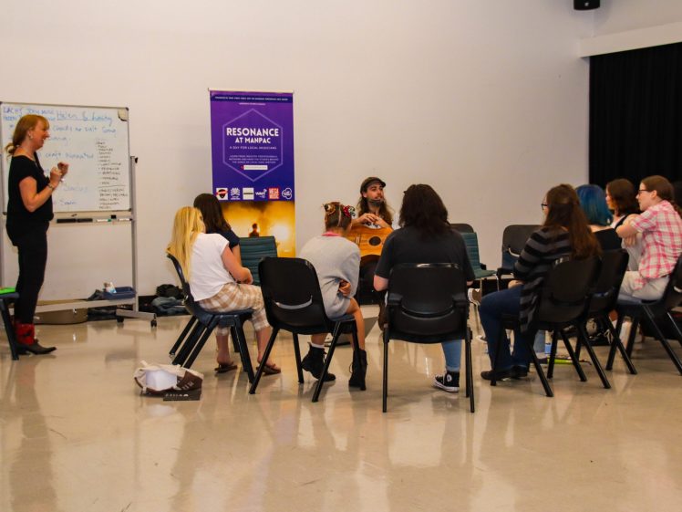 Image of A group of people sit in a circle of chairs at a workshop. One person stands by a whiteboard, whilst another faces the group with a drum. A Resonance at MAPAC banner is visible on the wall.
