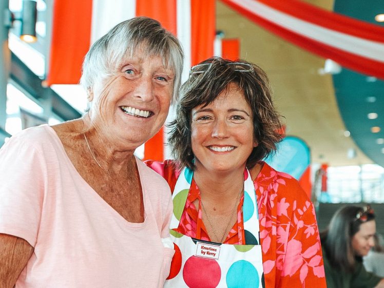 Image of Two women smiling at the camera; one wears a light pink shirt, the other wears a colourful polka-dot apron over a red floral top. They are indoors with festive red and white decorations in the background.