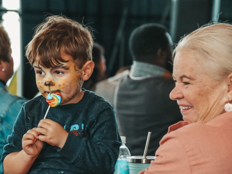 Image of A young child with face paint resembling a dog licks a colourful lollipop while an older woman with white hair, wearing a peach top, smiles beside him. Other people are blurred in the background.