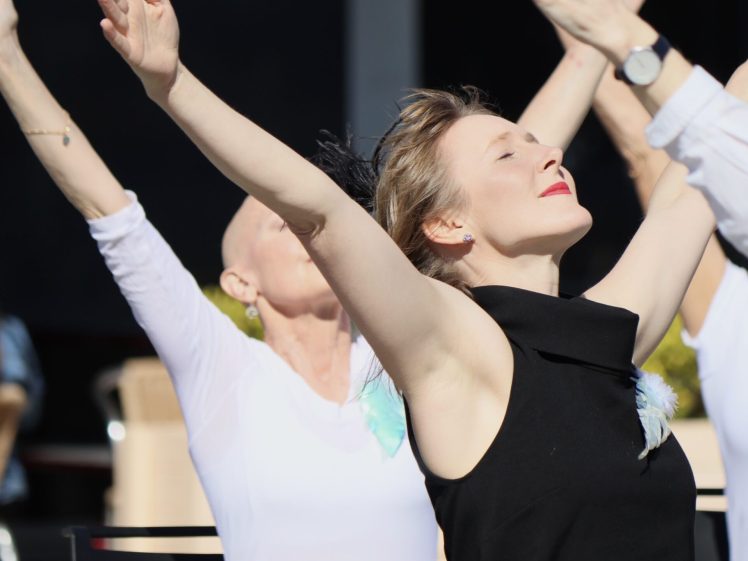 Image of A woman in a black sleeveless top stands with arms raised and eyes closed, facing the sun, surrounded by others with arms raised in a similar pose, suggesting a moment of joy or group exercise outdoors.