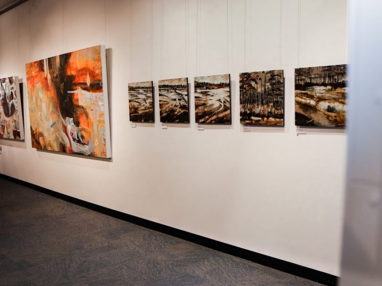 Image of A gallery wall displays several abstract and landscape paintings of different sizes. The artworks hang neatly in a row on a white wall above a grey carpeted floor.