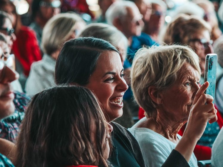 Image of A group of people sit closely together at an indoor event. A woman in the middle is smiling and laughing, while another woman next to her holds up a mobile, appearing to take a photo or video. The atmosphere looks lively and cheerful.