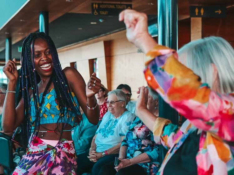 Image of A smiling woman with long plaits dances joyfully with another woman in a colourful outfit, while a group of older seated people watch and smile in the background.