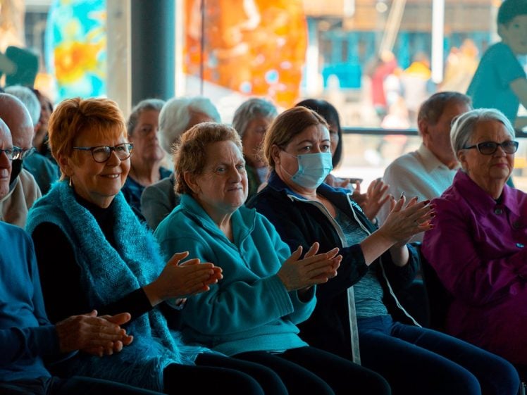 Image of A group of older adults sit closely together in rows, clapping and smiling. One woman in the front wears a face mask. The atmosphere appears cheerful and supportive.
