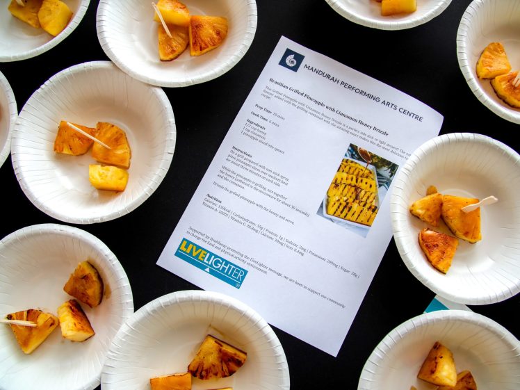 Image of Paper plates with grilled pineapple pieces on cocktail sticks surround a printed recipe for grilled pineapple on a table. The recipe sheet has a photo of the dish and logos for Mandurah Performing Arts Centre and LiveLighter.