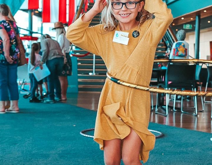 Image of A young girl in a yellow dress and glasses hula hoops indoors at an event. She smiles at the camera, with people and a LiveLighter banner in the background.