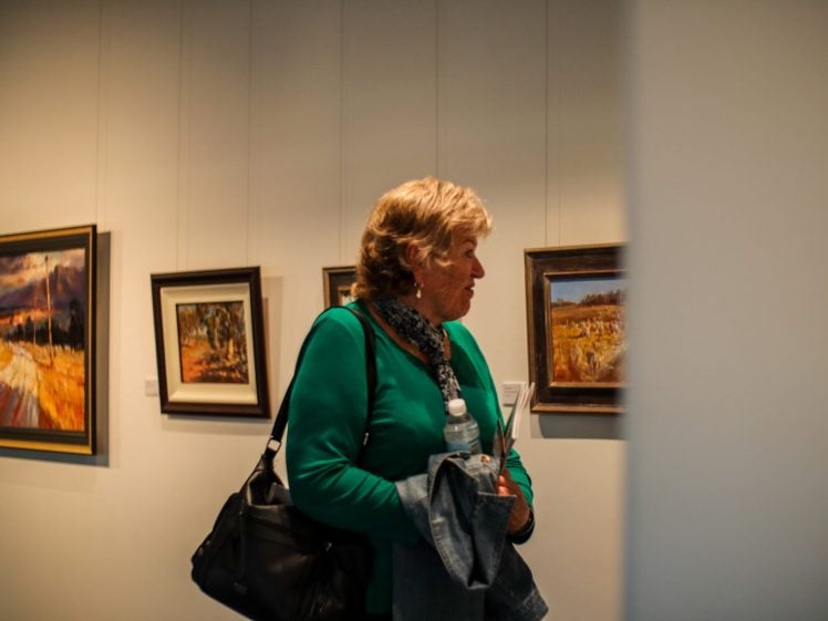 Image of A woman in a green top stands in an art gallery holding a jacket, water bottle, and papers, with framed landscape paintings hanging on the wall behind her.